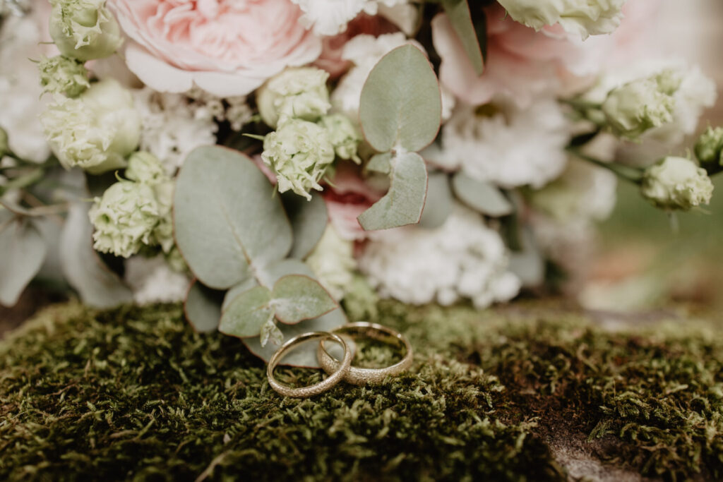 Eheringe im Moos vor einem Brautstrauß mit rosa Blumen
