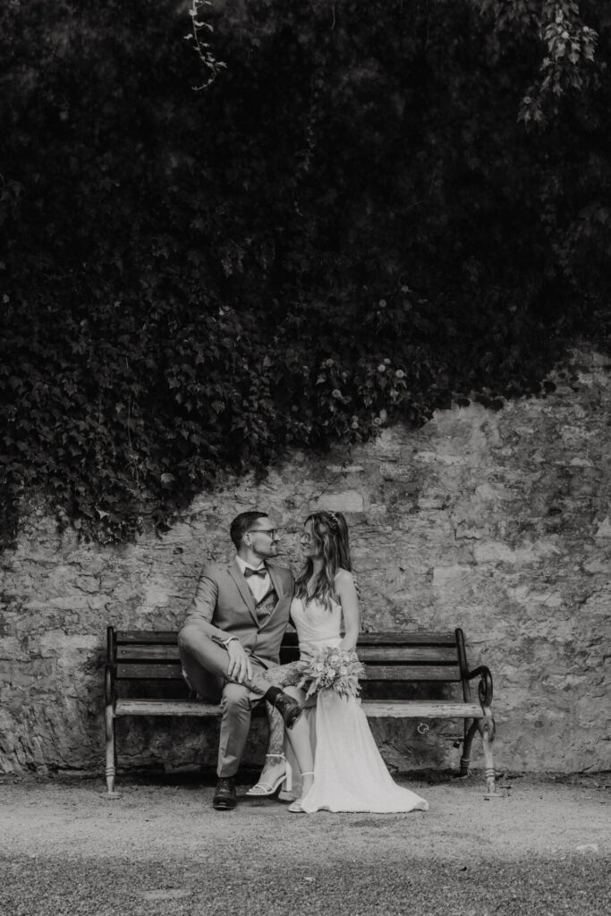 Brautpaar sitzt auf einer Bank vor einer alten Steinwand