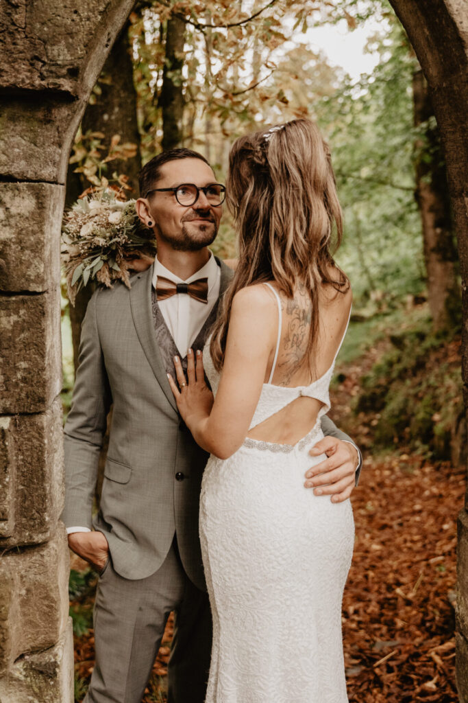 Braut und Bräutigam stehen eng unter einem Steinbogen im Wald bei ihrer Hochzeit