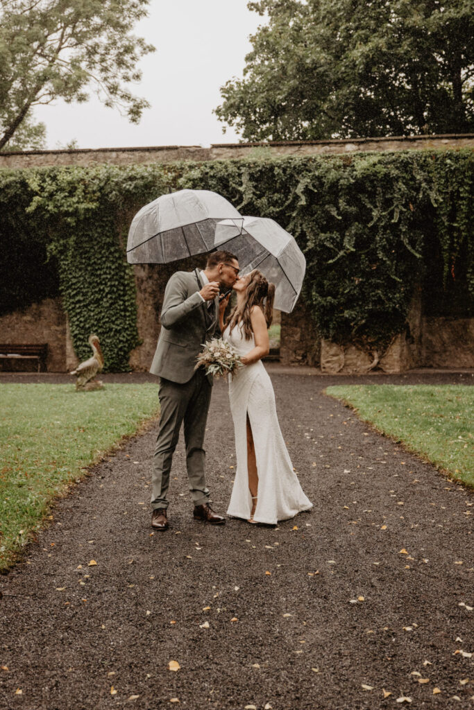 Brautpaar küsst sich im Regen unter transparenten Schirmen